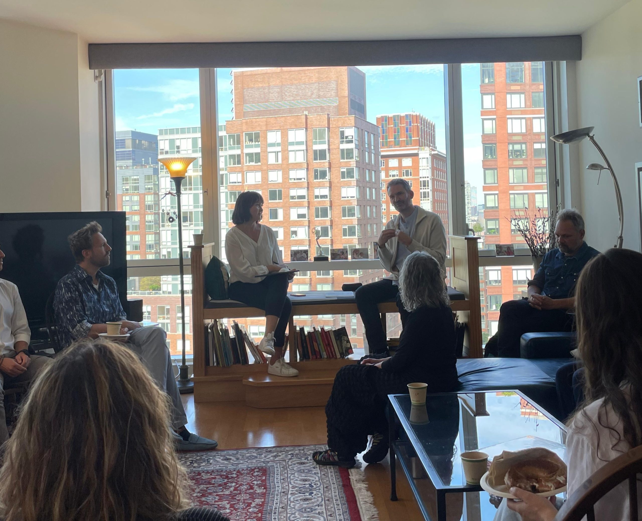Guests seated in a circle at a book launch, a city skyline through the window behind them