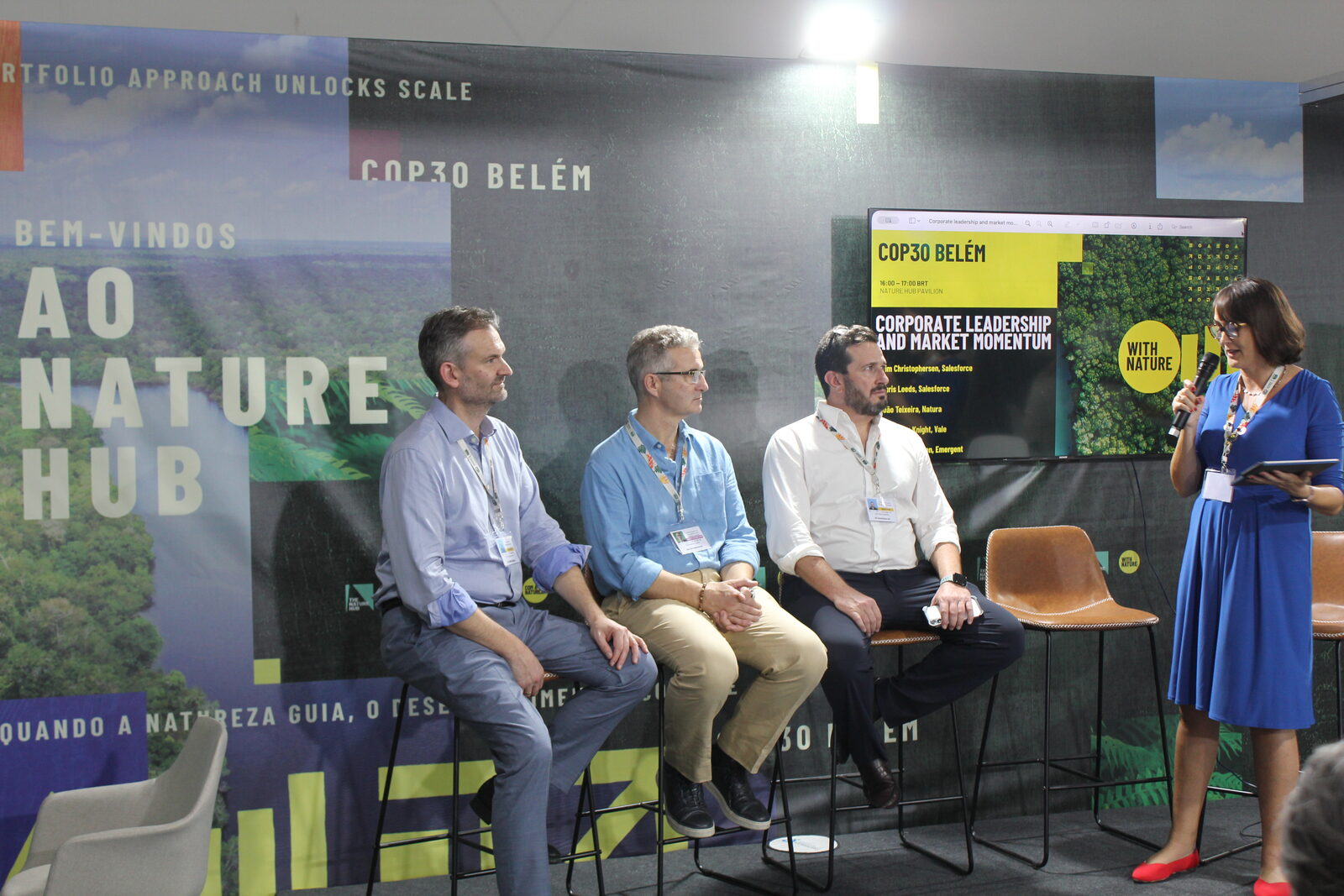 Jen Stebbing with a microphone addressing three seated panellists at the Nature Hub Pavilion, COP30 Belem