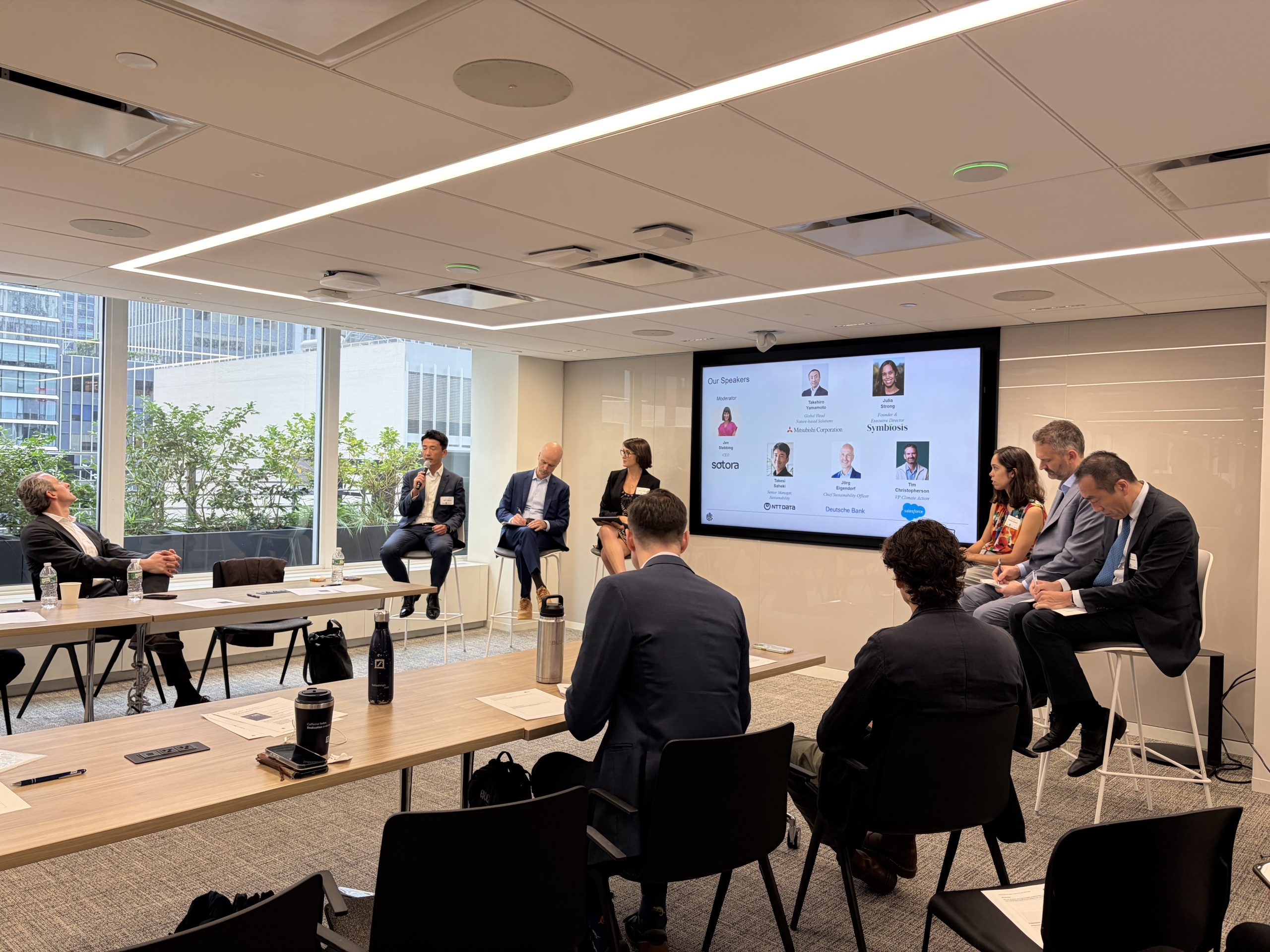 Speakers seated at the front of a boardroom session at Climate Week NYC, beside a screen showing panellist photos